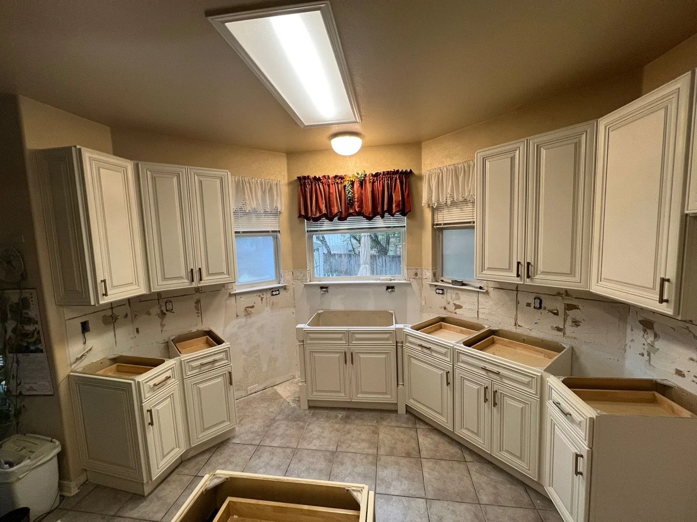 New cream raised-panel kitchen cabinets set on an existing tile floor, old backsplash scraped off the wall, countertops not yet installed